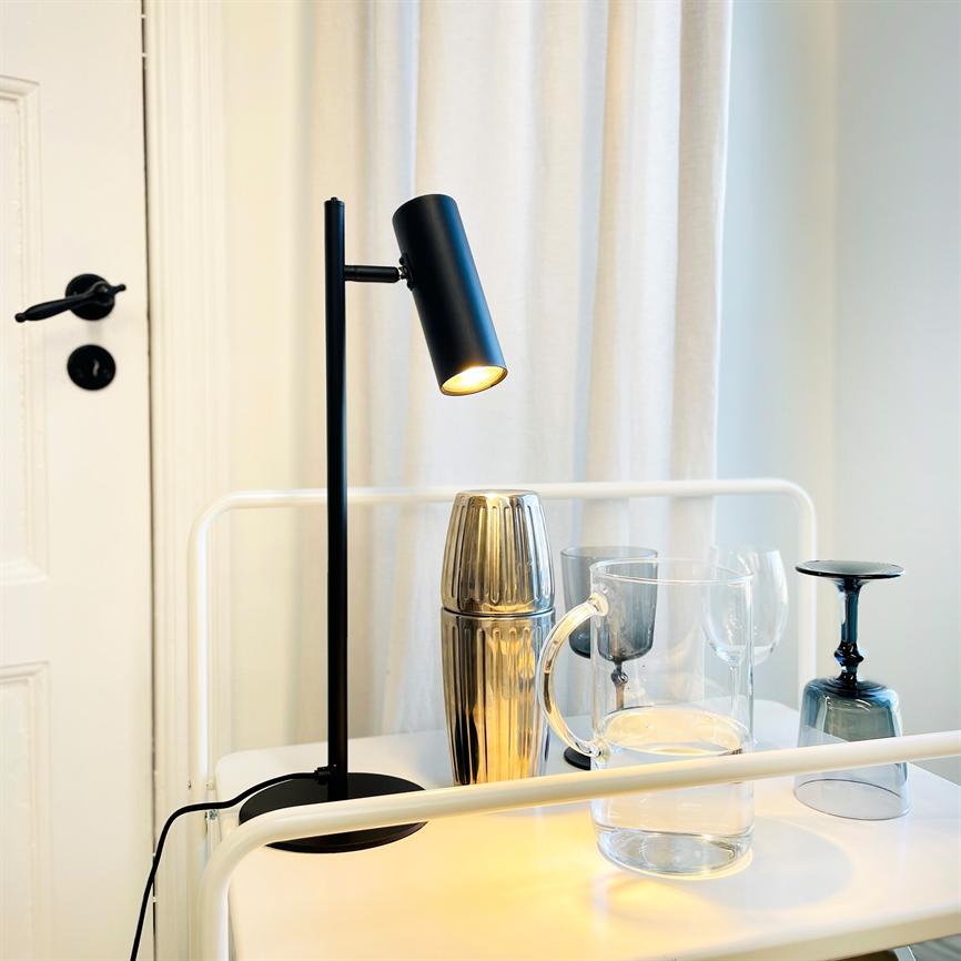 Sort bordlampe lyser ned på en glas kande og en blank cocktailshaker, omgivet af oprejste vinglas på en hvid rullevogn foran lyse gardiner og en hvid dør.