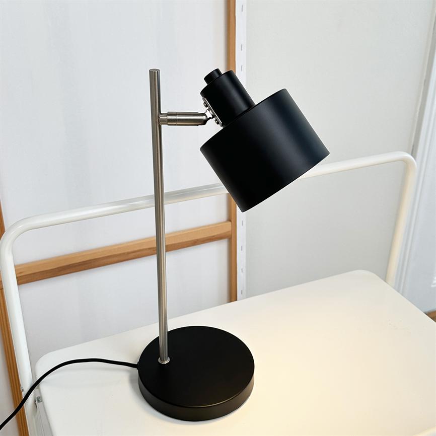 Sort skrivebordslampe står på et hvidt bord; skærmen er vinklet og kaster lys nedad. Lampen har en sølvfarvet stang og rund sort fod, foran en hvid væg med træramme.