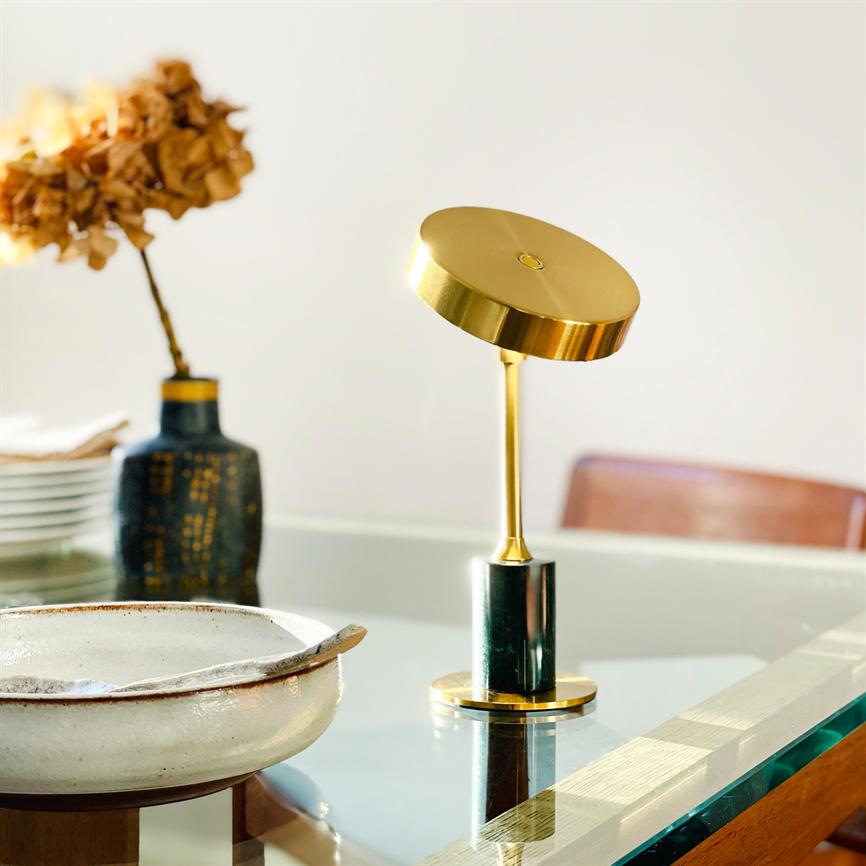 Guld bordlampe vipper i en skrå vinkel, placeret på et glasbord; foran en rustik keramikskål og ved siden af en mørk vase med tørrede blomster i et lyst spiserum.