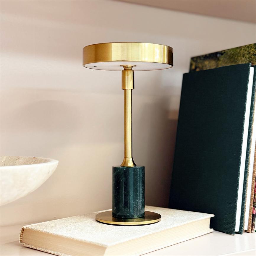 En bordlampe står på en indbundet bog på en hylde, ved siden af grønne bøger og en hvid skål; guldmetalisk skærm med mørk marmorfod.