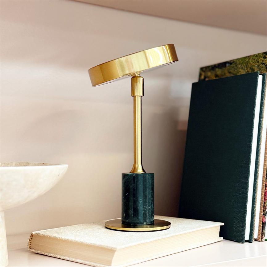 Læselampe i børstet messing, med skråtstillet lampehoved, står på en mørkegrøn marmorfod placeret oven på en lukket bog på en hylde, flankeret af to mørke indbundne bøger.