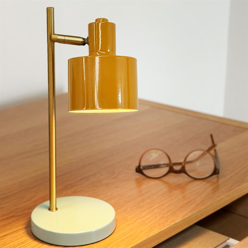 En gul metal bordlampe lyser nedad over et træskrivebord; en rund lys base og slank messingstang står ved siden af et par runde briller.