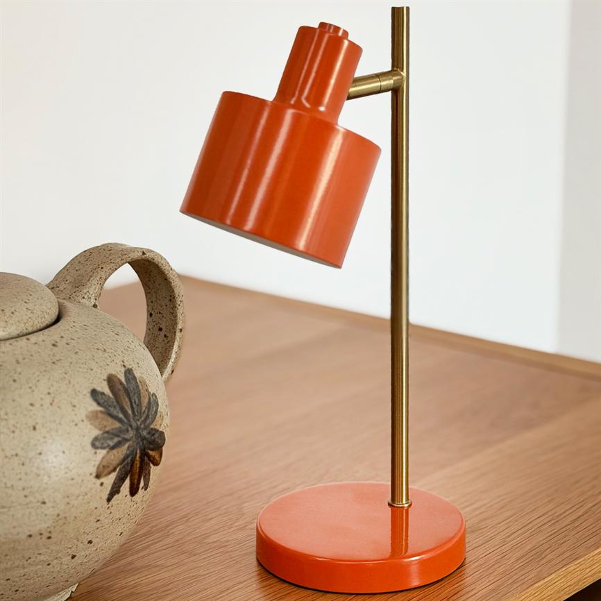 Orange bordlampe står på en slank messingstang og vipper nedad over et træbord ved siden af en beige, prikket keramikvase med sort blomsterdekoration foran en lys væg.