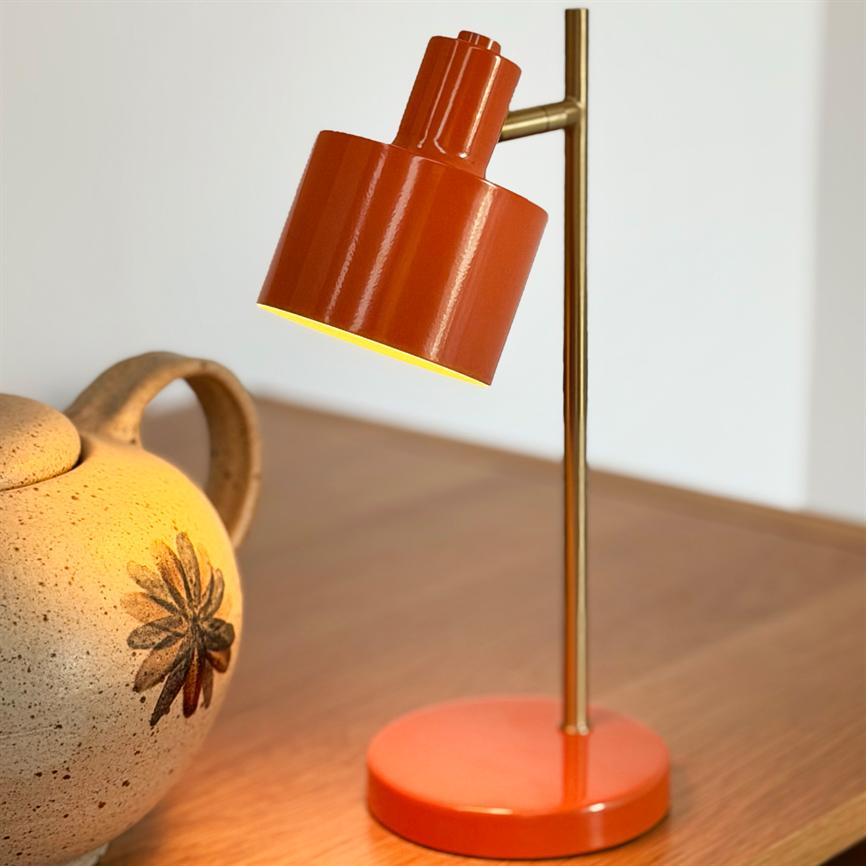 Rød-orange bordlampe kaster varmt lys nedad; slank messingstang holder skærmen over et træbord ved siden af en beige, dekoreret keramisk kande.