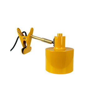Klemmelampe med metalarm klemmer fast via sort ledning og gummibelagte kæber, understøtter en cylindrisk gul lampefod; produktfoto på ensartet hvid baggrund.