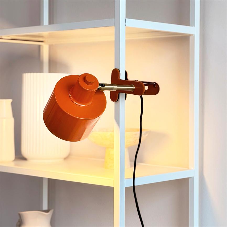 En orange klemme-lampe sidder fast på en hvid metallehylde og lyser nedad; hylden rummer hvide keramiske vaser og en lys skål i et varmt, blødt lys.