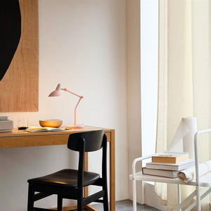 Rosa skrivebordslampe kaster blødt lys over en åben notesbog og en skål på et træskrivebord; omkring står en sort stol, lyse gardiner og en hvid hylde med bøger.