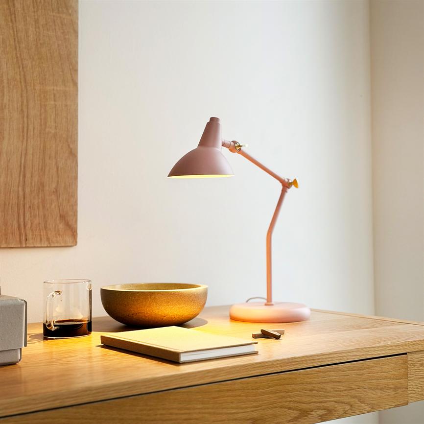 En rosa skrivebordslampe kaster varmt lys over et egetræsbord med en keramisk skål, notesbog, glas med kaffe og småclips i et lyst, minimalistisk værelse.