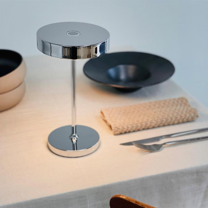 Krombordlampe står midt på et hvidt dækket bord og reflekterer omgivelserne; omkring ligger en sort dyb tallerken, beige skåle, et sammenfoldet sandfarvet viskestykke og bestik.