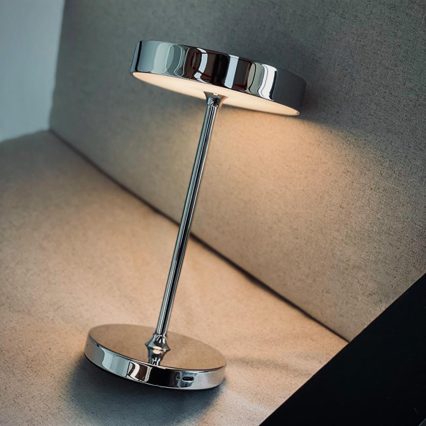 En krom bordlampe lyser blødt nedad, hældende i en vinkel; lampen står på sofaens beige polstrede sæde mod ryglænet med reflekterende metalbase og rund skærm.