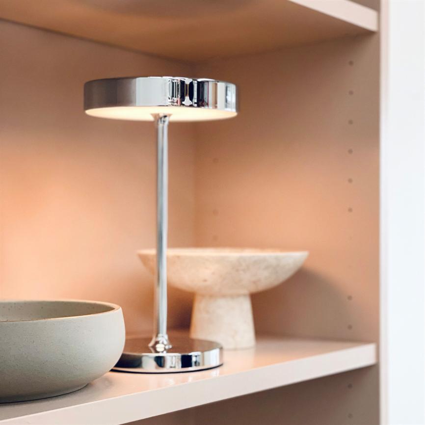 En krom bordlampe kaster blødt lys på en lyserosa hylde ved siden af en rund grå skål og en fodet beige skål.