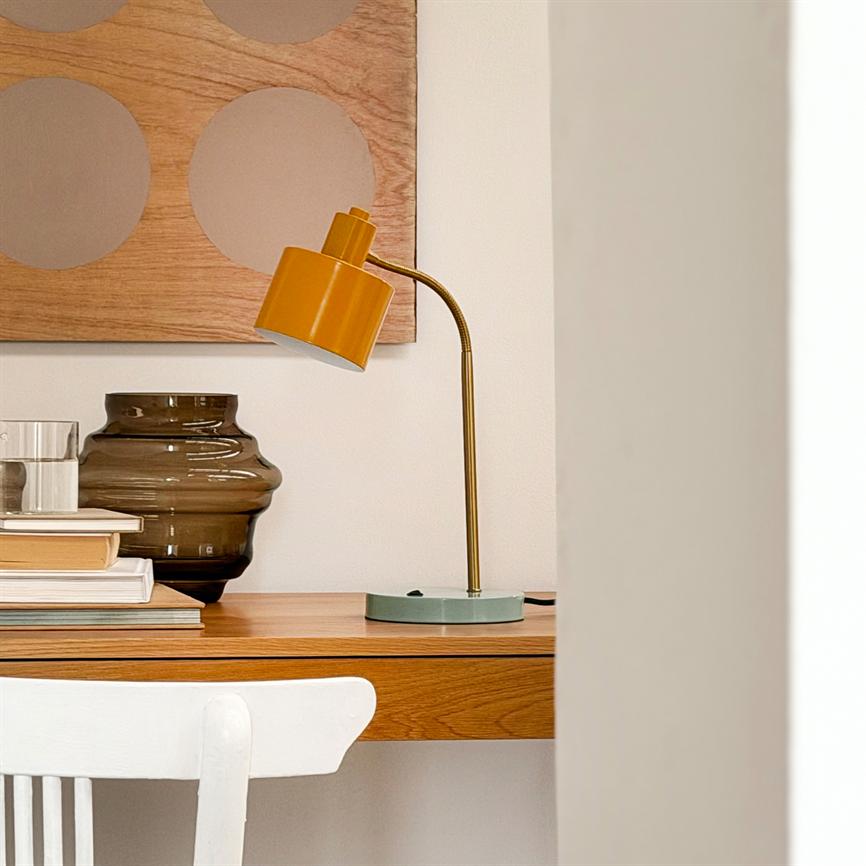 Gul skrivebordslampe bøjer sig over et træbord; ved siden står en brun glasvase og en stak bøger, foran en hvid stol og et cirkelmønstret træpanel på væggen.