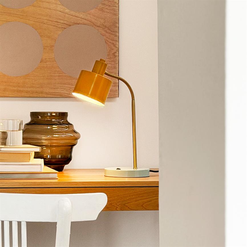 Gul skrivebordslampe lyser på et træbord, flankeret af en brun glasvase, bunker af bøger og et abstrakt træpanel på væggen, med en hvid stol foran bordet.