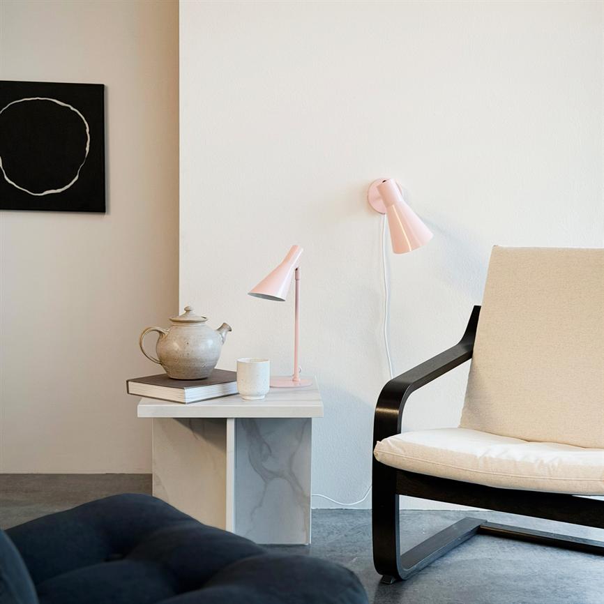 En beige lænestol står ved et lille marmorbord; bordet bærer tekande, krus og en lyserød skrivebordslampe, mens en lyserød væglampe og et sort cirkelmaleri hænger på væggen.