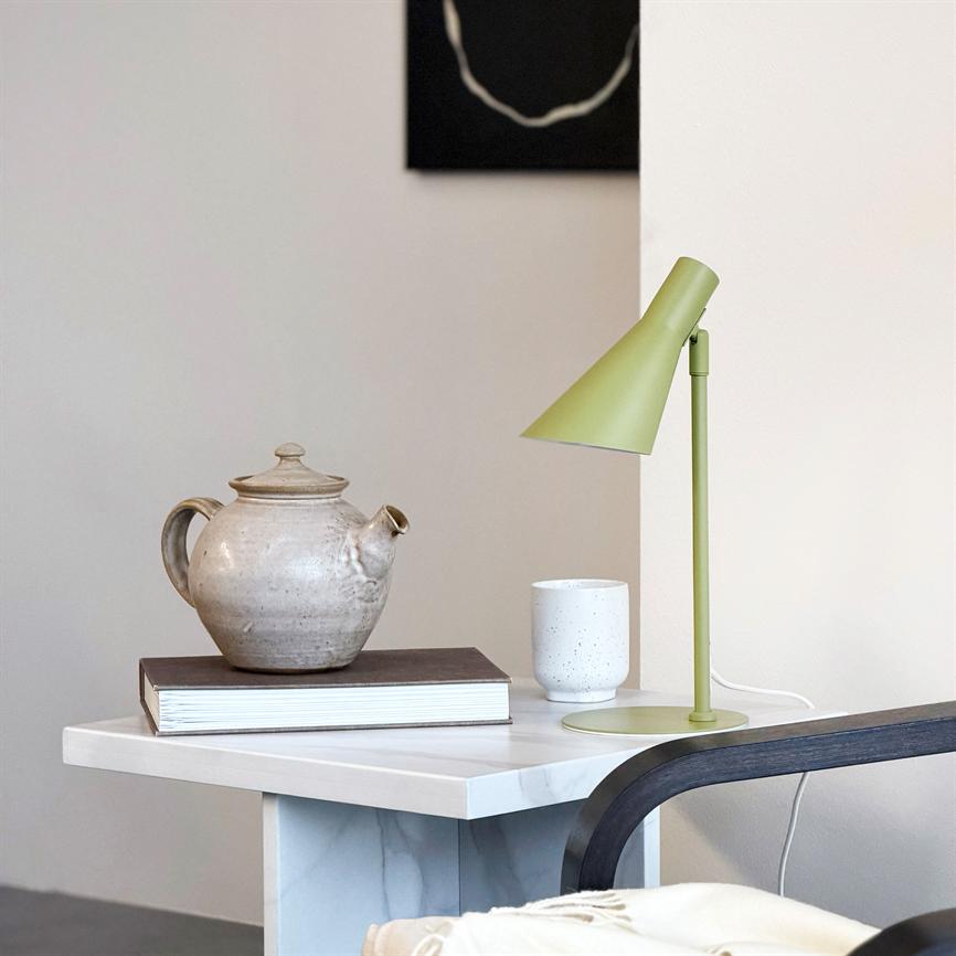 En beige keramisk tekande hviler på en lukket bog på et hvidt sidebord; en olivengrøn bordlampe står ved siden af sammen med en hvid kop i et lyst, minimalistisk rum.