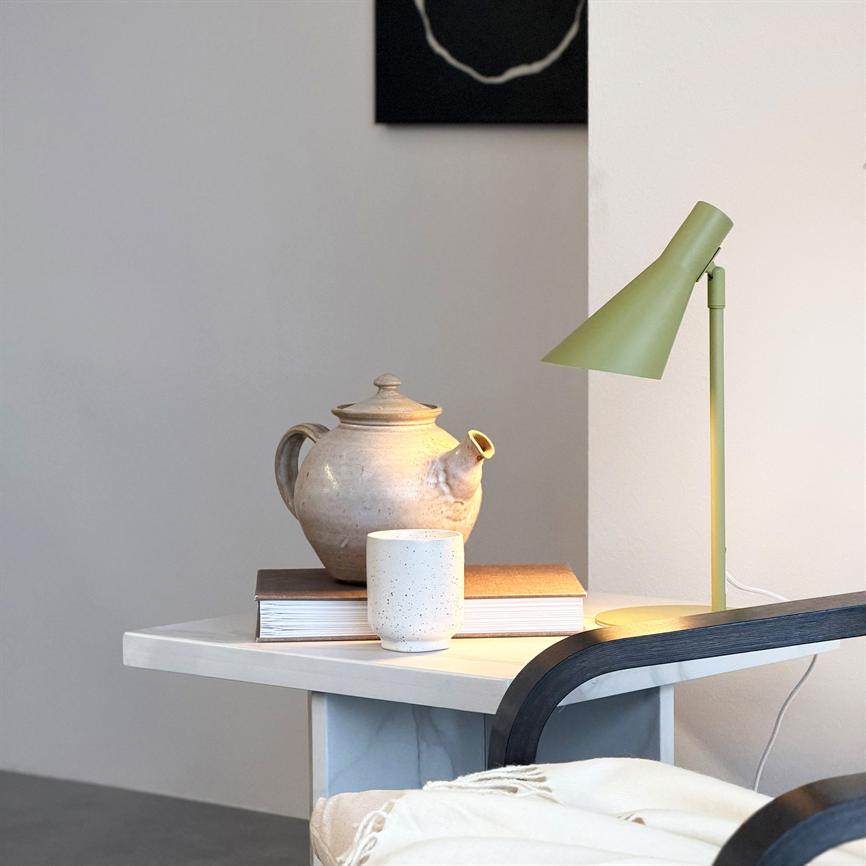 En lertekande hviler på en stabel bøger, en keramikkop står foran den; en grøn bordlampe kaster varmt lys over genstandene på et hvidt sofabord i et roligt hjørne.