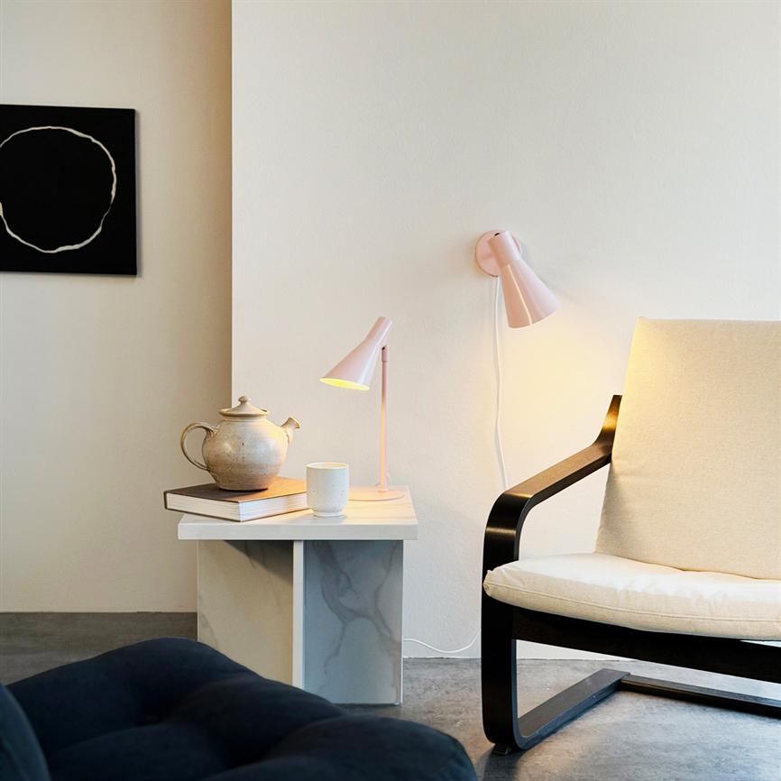 Lænestol står ved et sidebord; bordet bærer tekande, kop og bog, en bordlampe lyser, og en væglampe kaster varmt skær i et lyst, minimalistisk rum.