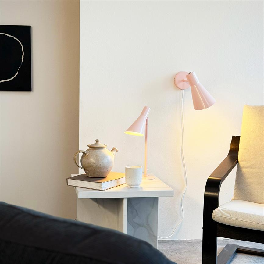 En keramisk tekande står på et sidebord, ledsaget af en kop og to lyserøde lamper, ved en lys væg og en lænestol med sorte armlæn i et roligt hjørne.