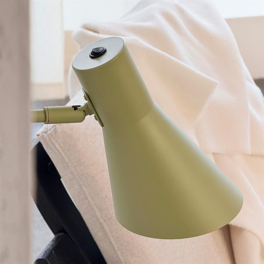 Grøn skrivebordslampe peger nedad fra en justerbar arm; den står nær en lysegrå lænestol med en krøllet cremefarvet plaid i et lyst, minimalistisk rum.