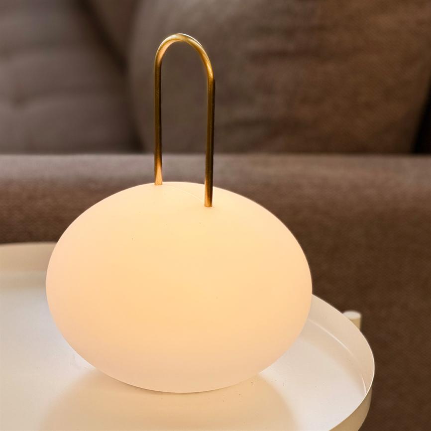 Rund bordlampe med gyldent bøjlehåndtag lyser blødt og varmt på en hvid bakke, foran en brun sofa i en stue.