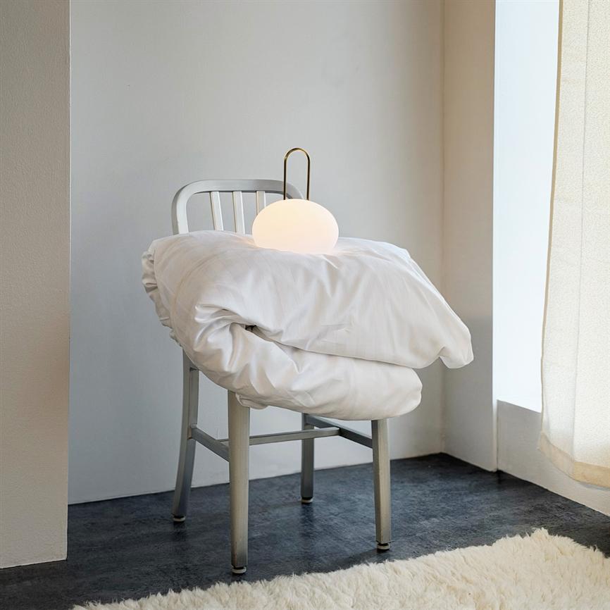 En metalstol bærer en foldet hvid dyne, hvorpå en rund, tændt lampe står; hjørnet er lyst med hvide vægge, mørkt gulv, lyst tæppe og sollys gennem et gardin.