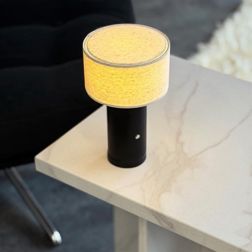 En lille bordlampe med cylindrisk, sort fod og beige stofskærm lyser varmt; den står på et lyst marmorbord ved en mørk lænestol og et lyst tæppe i et moderne rum.