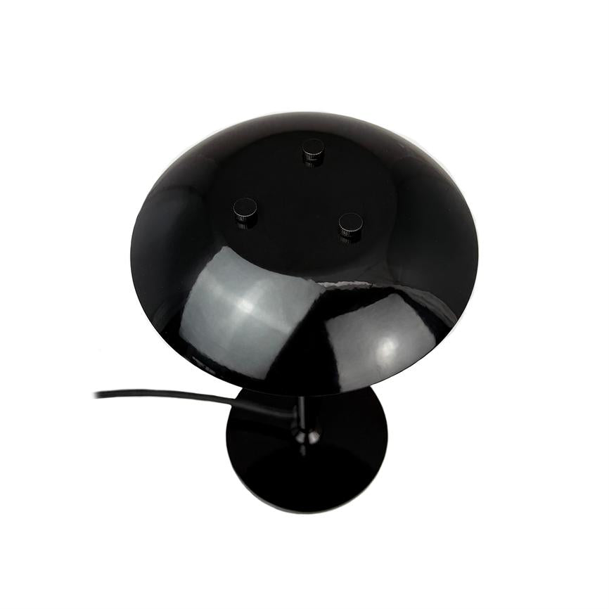 Sort bordlampe med rund, skålformet skærm og tre små knapper på toppen, med synlig ledning; skærmen reflekterer stærkt lys på en neutral, hvid baggrund.