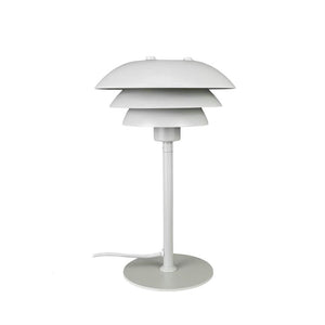 Hvid bordlampe med tre overlappende, skiveformede skærme, står på en slank stang og rund fod; tekstilledning ligger ved siden af, placeret mod ensartet hvid baggrund.