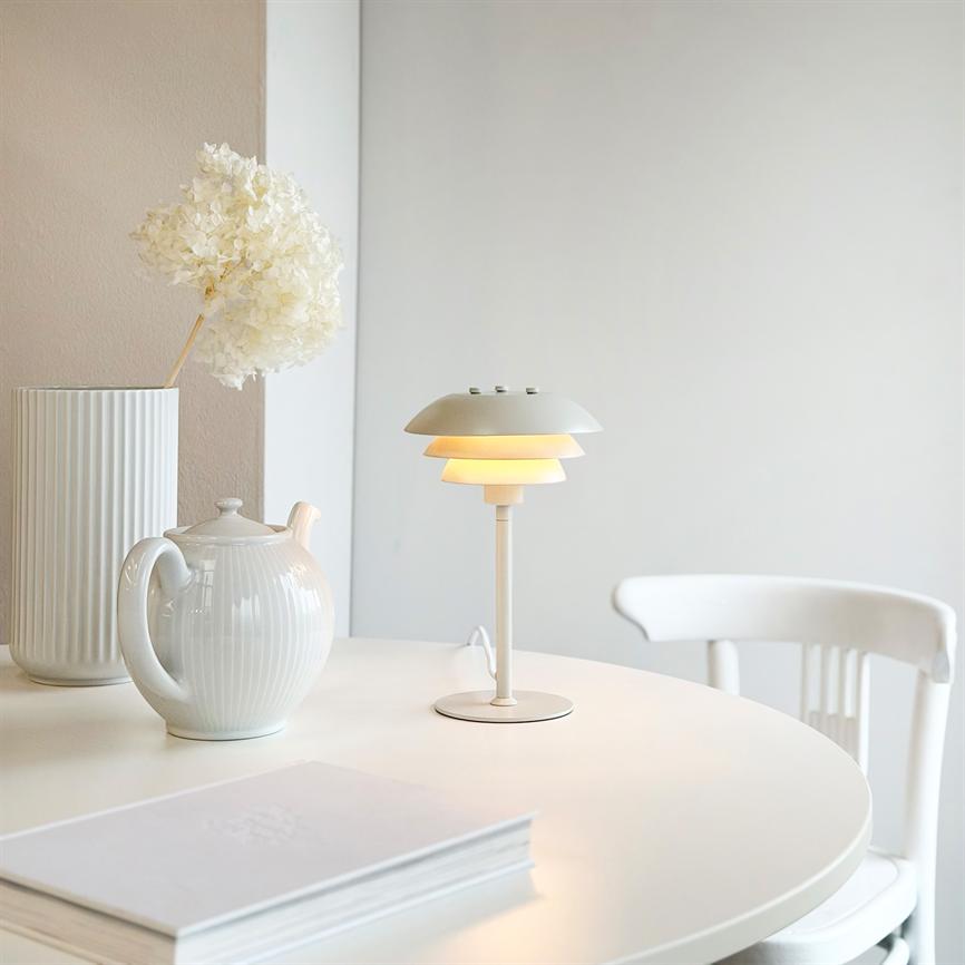 Hvid bordlampe udstråler blødt, varmt lys på et rundt hvidt bord; ved siden står en porcelæns tekande, en høj, rillet vase med tørret blomst og en hvid stol.