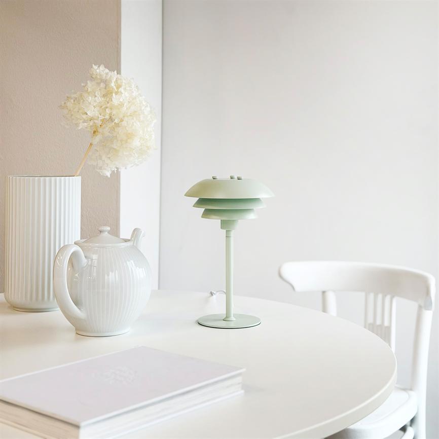 Lille mintgrøn bordlampe står midt på et rundt hvidt bord, omgivet af en hvid tekande, en høj, rillede vase med tørret blomst og en bog i et lyst, minimalistisk rum.
