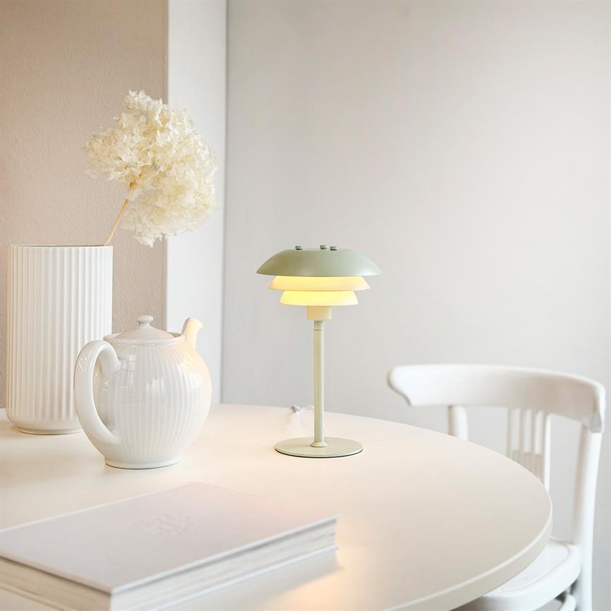 Bordlampe står tændt på et rundt hvidt bord omgivet af en hvid tekande, en høj vase med riller og en hvid træstol i et lyst, minimalistisk rum.