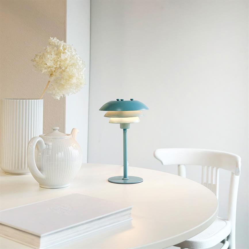 En turkis bordlampe står tændt på et rundt hvidt bord; ved siden af står en hvid tekanne og en høj vase med tørret blomst, med en hvid stol i baggrunden.