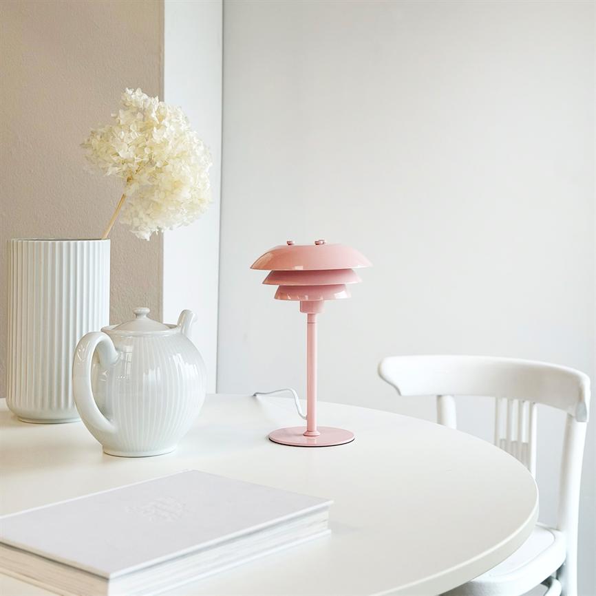 Lyserød bordlampe står på rundt hvidt bord; ved siden af en hvid tekande og vase med tørrede hvide blomster, foran en hvid stol og en neutral væg.