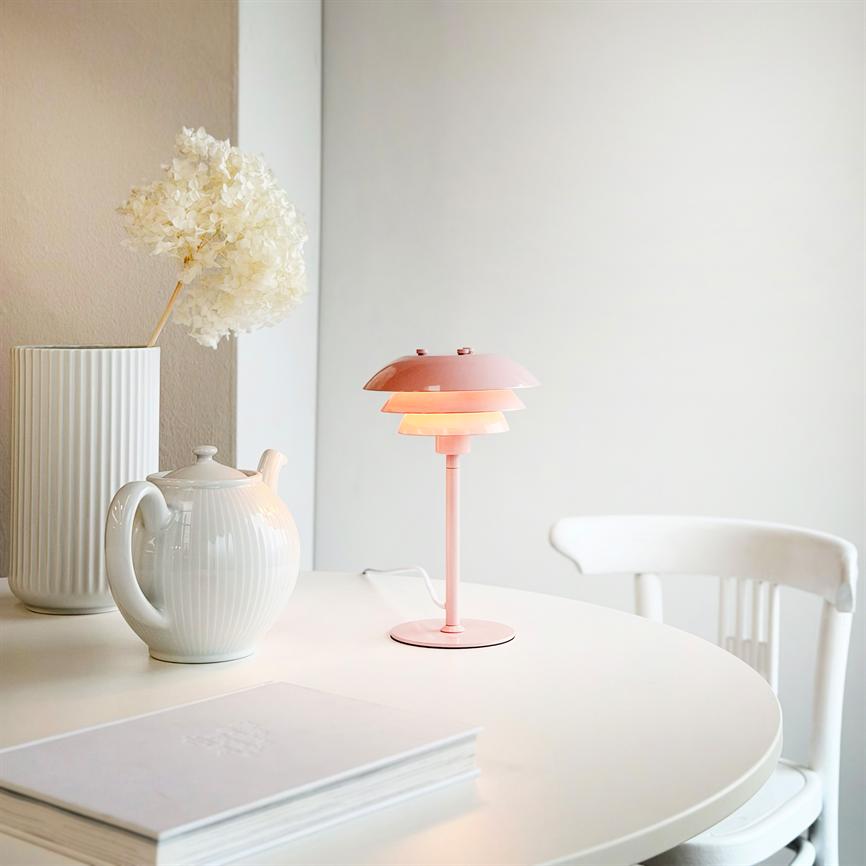 En lyserød bordlampe lyser varmt på et rundt hvidt bord, omgivet af en hvid tekande, en høj vase med tørret blomst og en ledig hvid stol i et neutralt rum.