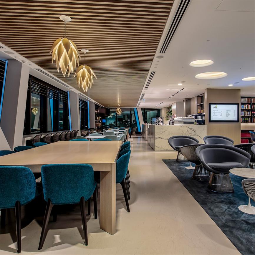 Et langt lyst træbord står centreret, omgivet af blågrønne stole; dekorative hængelamper lyser, lænestole og en marmorbænk-café i baggrunden, moderne lounge med træloft og store vinduer.
 
 Synlig tekst: "CAFÉ" (resten af skærmens tekst er ikke læselig).