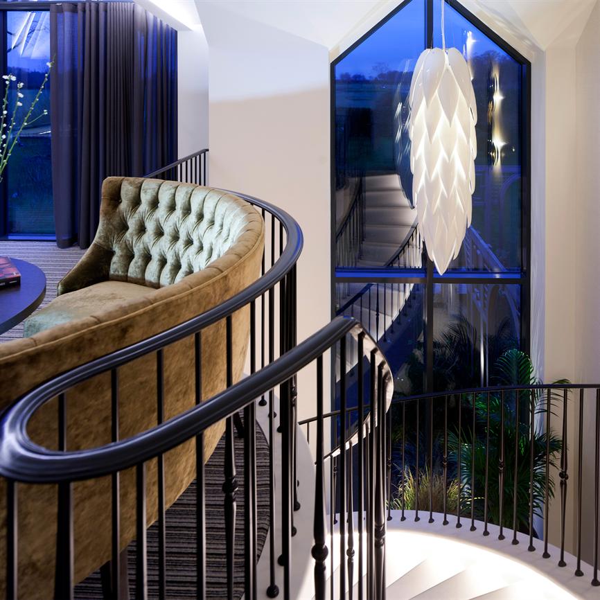 Et sort, buet gelænder omslutter en grøn fløjlssofa og fører ned ad en hvid, oplyst trappe; et stort trekantet vindue viser blå aftenhimmel og en hængende hvid dekorativ lampe.